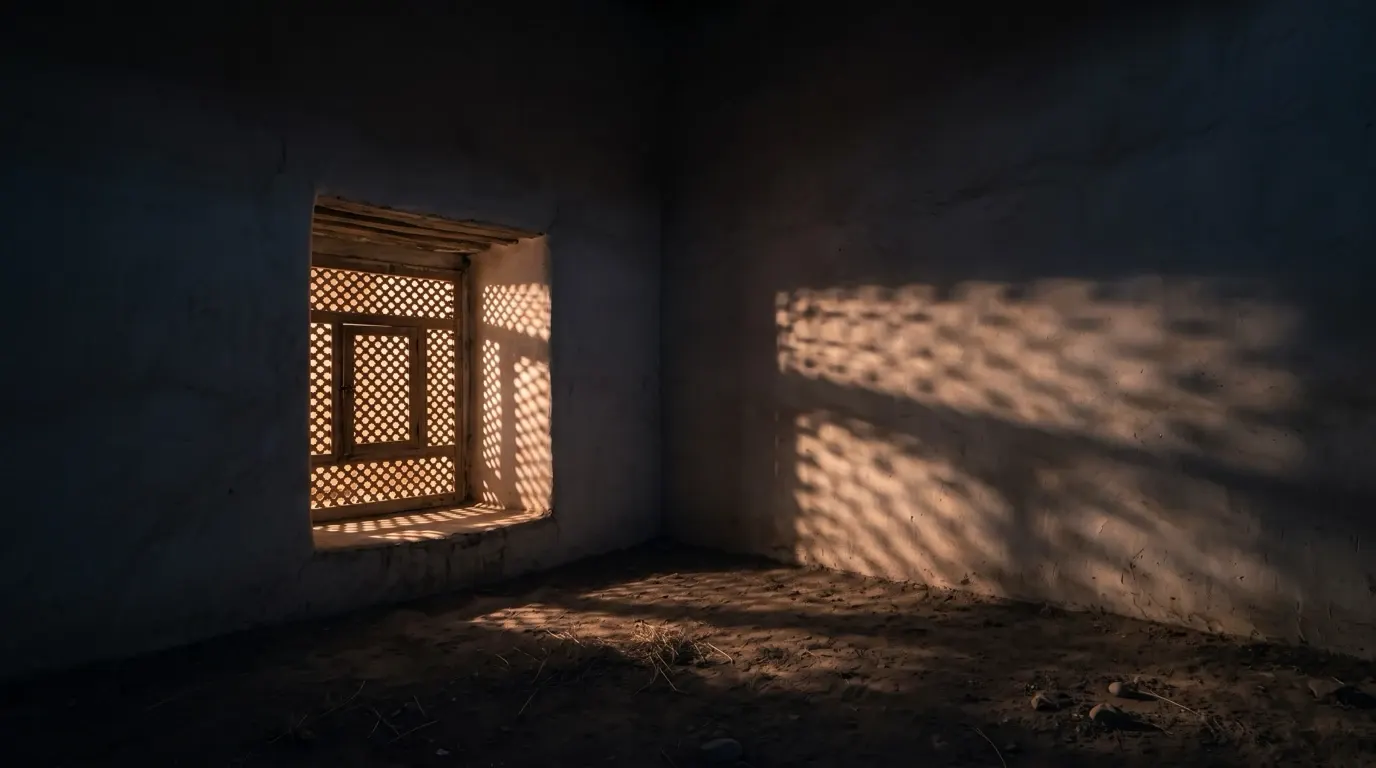Light falling through a wooden lattice onto a plastered wall and an earthen floor.
