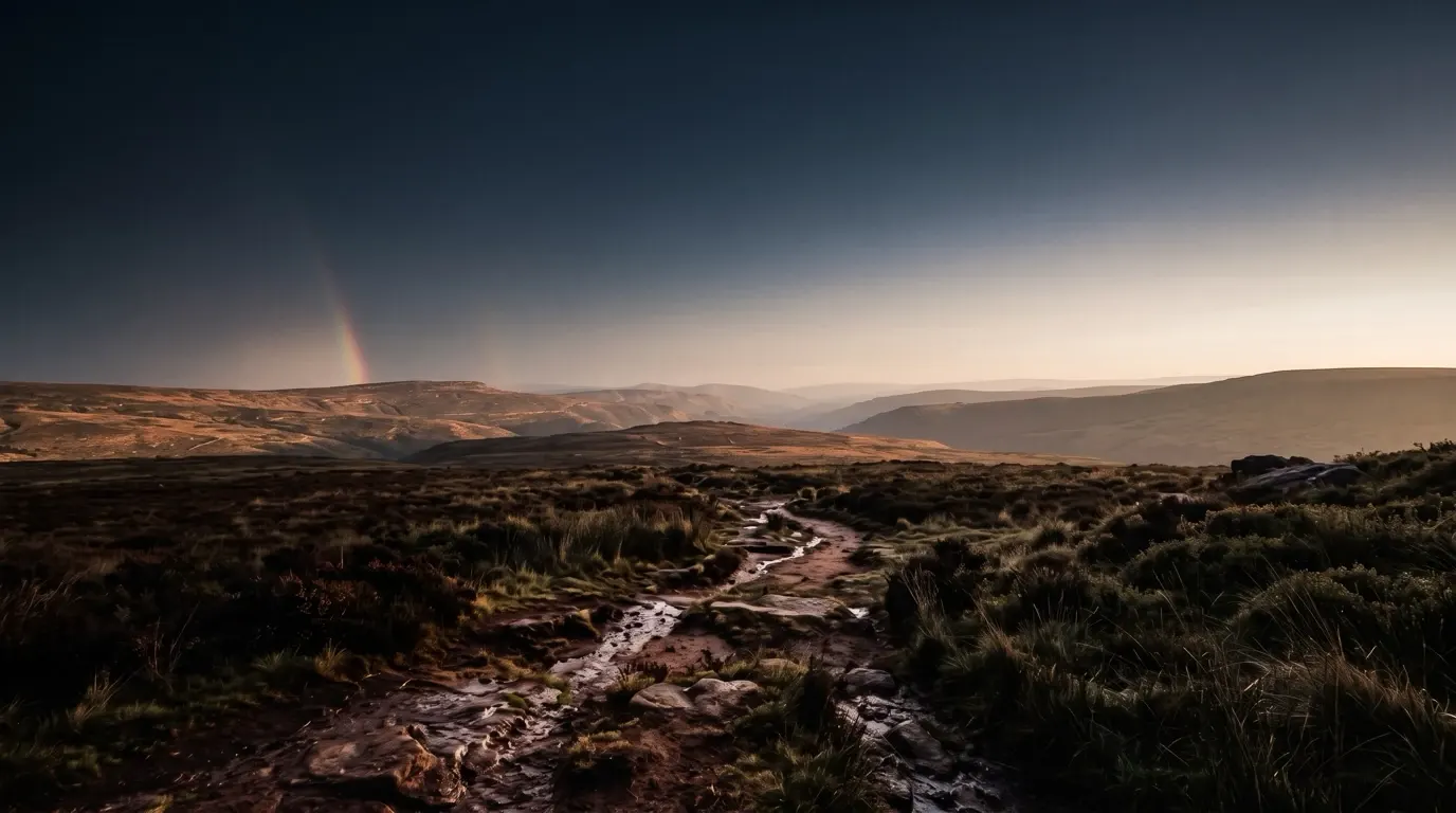 Wet hill country after long rain, a short fragment of a faint arc rising at the edge of the sky.