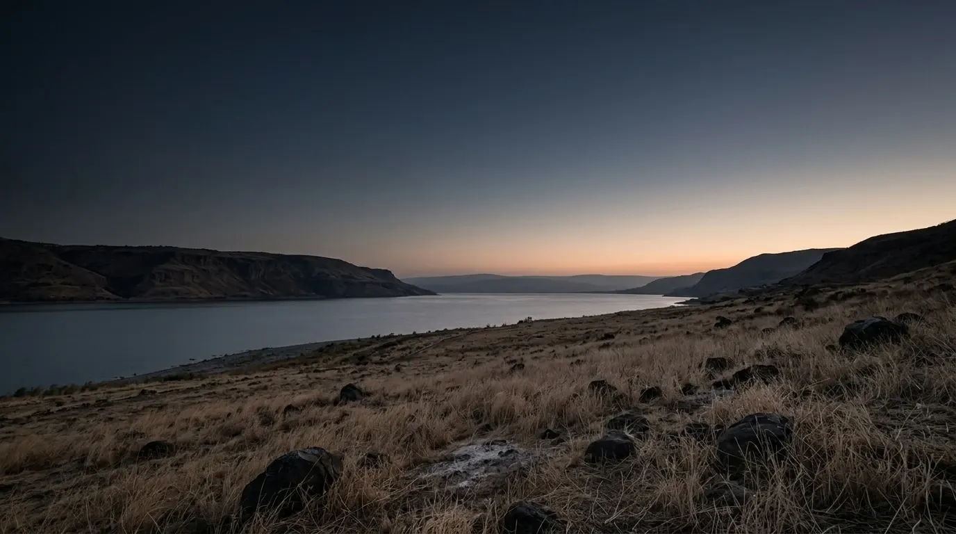 A wide slope of dry coarse grass running down to a broad still lake, dark basalt in the near ground.