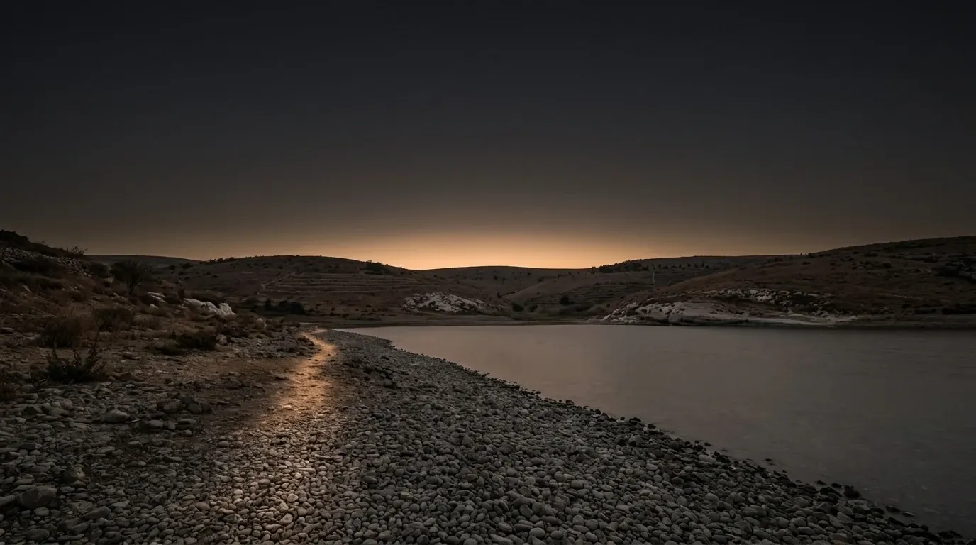 A shore of grey rounded pebbles running down into a wide still freshwater lake, low dry brown hills closing the far side.