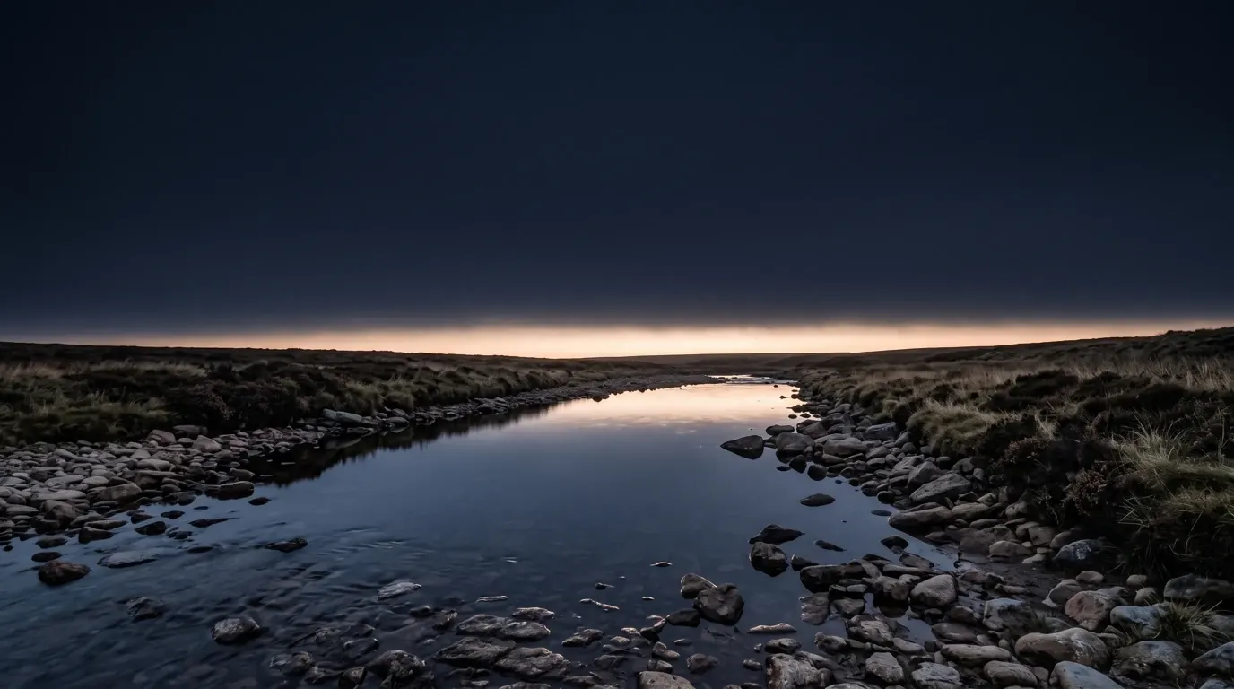 A shallow river running through open moorland, low stone banks and coarse grass, the water still.