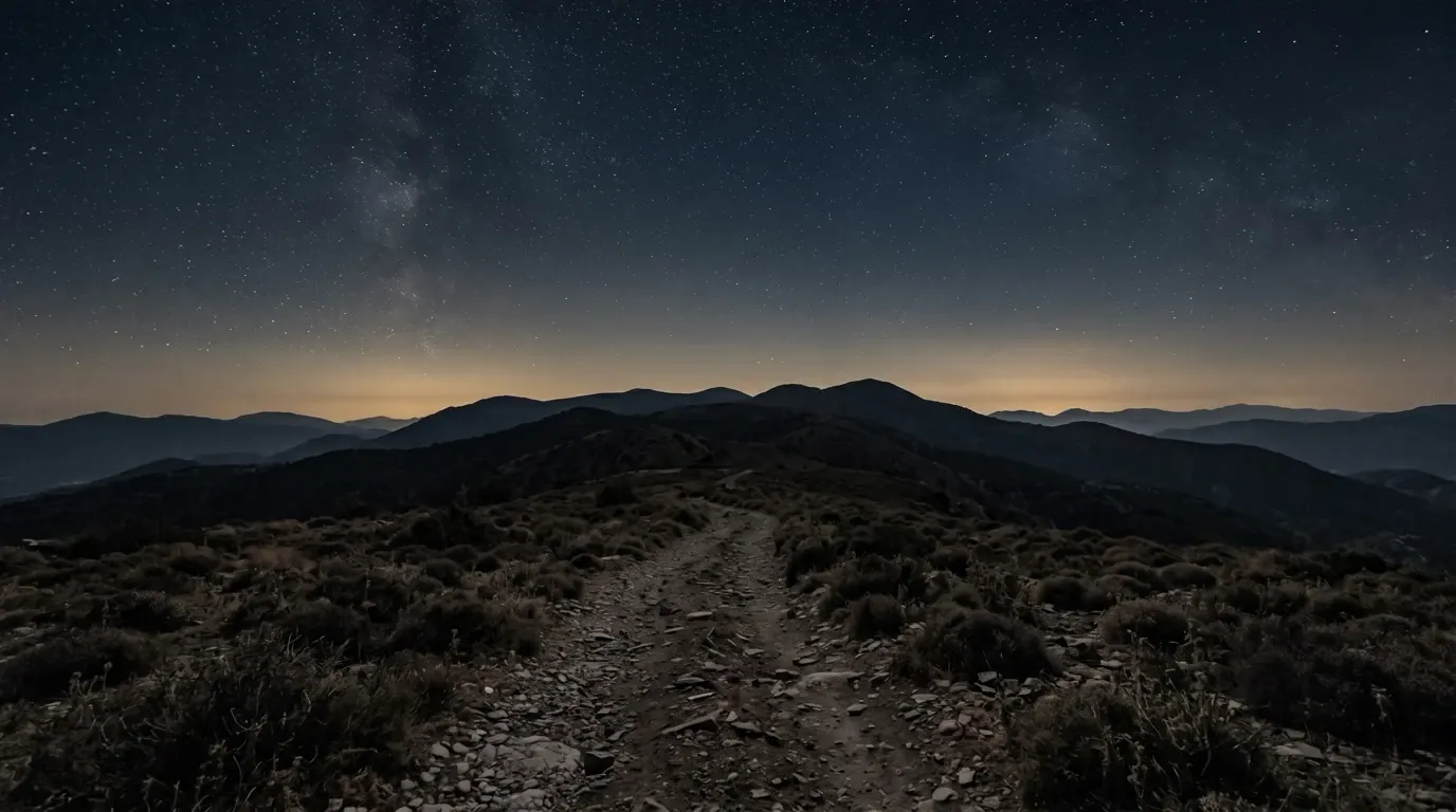 A deep sky full of stars over dark mountain ridges, a stony path in the foreground.